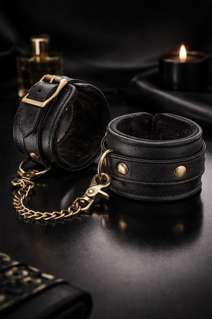Hochwertige schwarze Lederfesseln mit goldenen Metallelementen und Verbindungskette, inszeniert in einer dunklen, eleganten Wellness-Atmosphäre mit warmem Licht und luxuriöser Ausstrahlung
