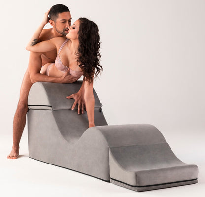 Liberator Aria Flip Chaise – graue Premium-Sexlounge in ausgeklappter, ergonomisch geschwungener Form, auf der eine erwachsene Frau in schwarzer Kleidung mit erhöht gelagertem Oberkörper und angewinkelten Beinen liegt – erhältlich bei Evoria Care