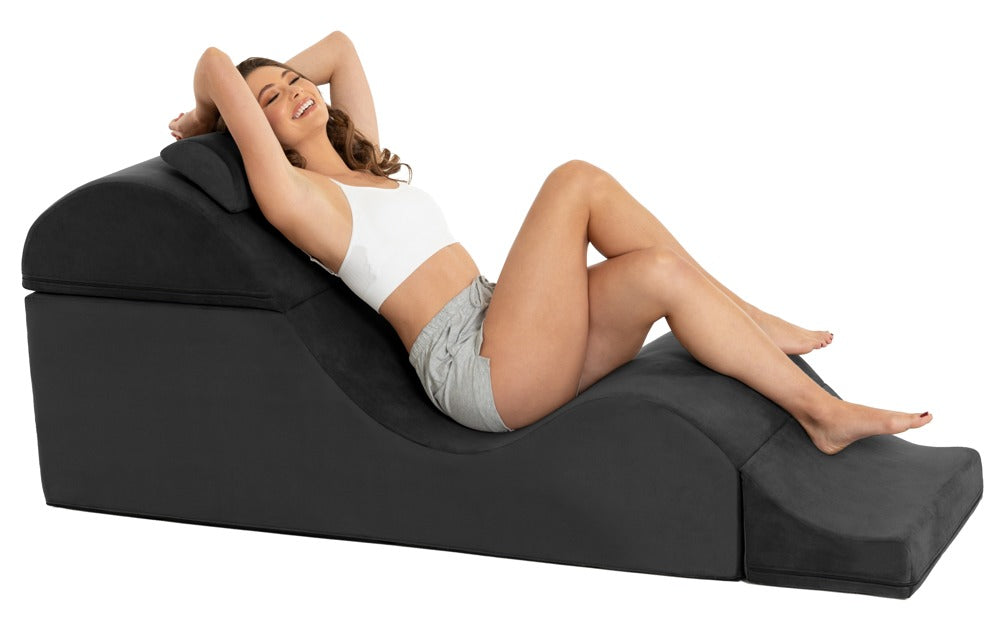 Liberator Aria Flip Chaise – schwarze Premium-Sexlounge mit geschwungener Polsterform, auf der eine erwachsene Frau mit erhöht gelagertem Oberkörper und angewinkelten Beinen liegt – erhältlich bei Evoria Care