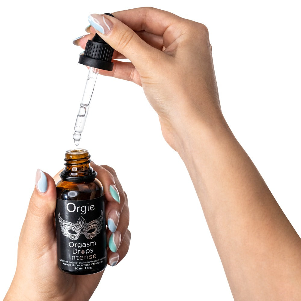 Orgie Orgasm Drops Intense wärmende Klitoristropfen in schwarzer Flasche mit herausgezogener Pipette, zwischen zwei Händen zur Veranschaulichung der Dosierung dargestellt – erhältlich bei Evoria Care