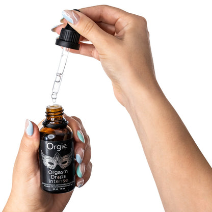 Orgie Orgasm Drops Intense wärmende Klitoristropfen in schwarzer Flasche mit herausgezogener Pipette, zwischen zwei Händen zur Veranschaulichung der Dosierung dargestellt – erhältlich bei Evoria Care