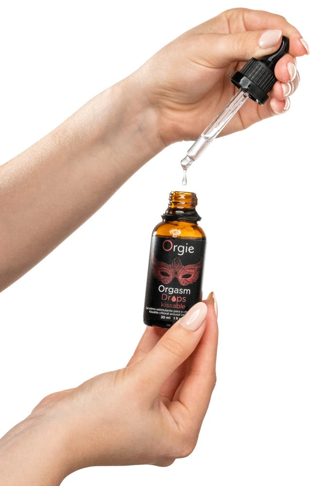 Orgie Orgasm Drops Kissable Klitoristropfen in schwarzer Flasche mit herausgezogener Pipette, zwischen zwei Händen zur Veranschaulichung der Dosierung dargestellt – erhältlich bei Evoria Care