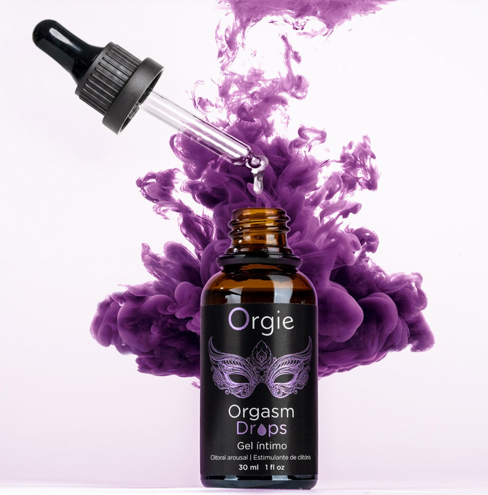 Orgie Orgasm Drops Klitoris-Tropfen in schwarzer Pipettenflasche mit violettem Etikett, dekorativ inszeniert mit violetten Blüten auf hellem Hintergrund – erhältlich bei Evoria Care