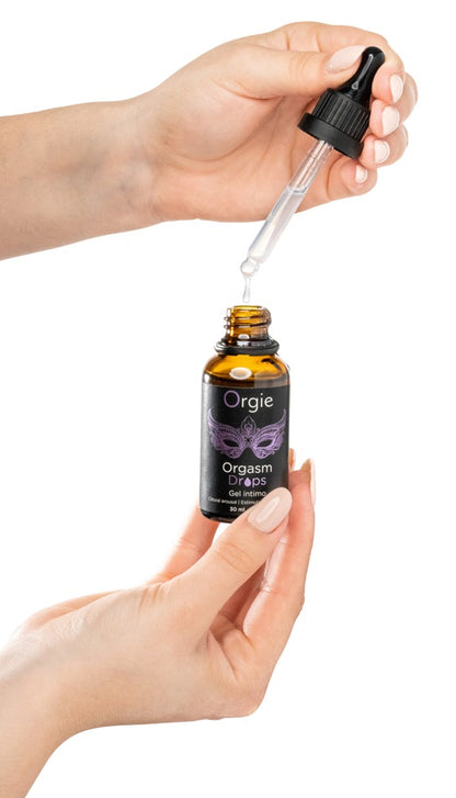 Orgie Orgasm Drops Klitoris-Tropfen in schwarzer Flasche mit herausgezogener Pipette, zwischen zwei Händen gehalten zur Veranschaulichung der Dosierung und Handhabung – erhältlich bei Evoria Care