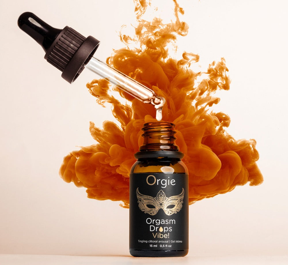 Orgie Orgasm Drops Vibe! Klitoristropfen in schwarzer Pipettenflasche mit goldfarbenen Akzenten, dekorativ inszeniert mit orangefarbenen Blüten und Blättern auf hellem Hintergrund – erhältlich bei Evoria Care